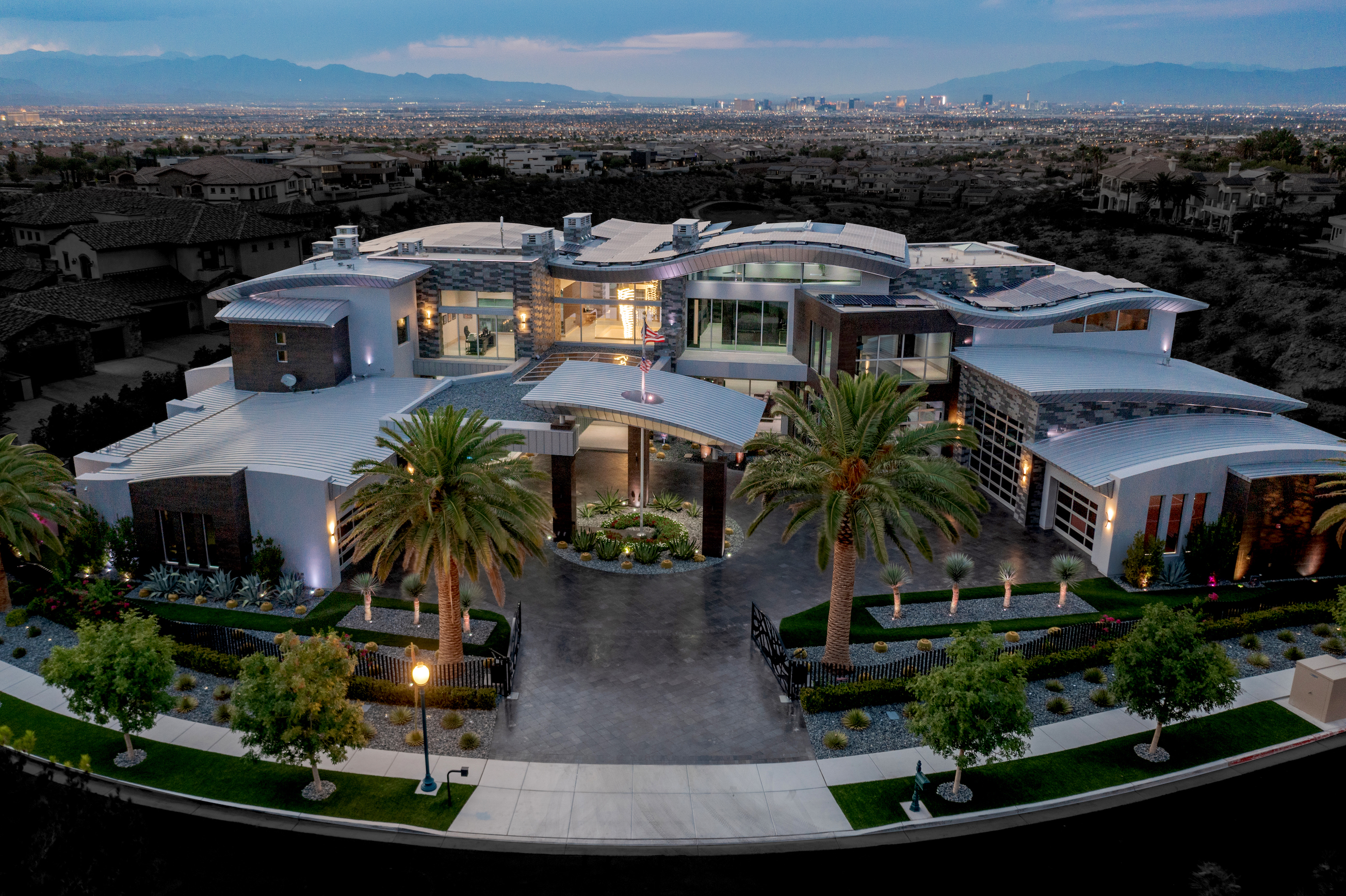 Expansive modern estate with solar-paneled roof set to Vegas strip backdrop at dusk.